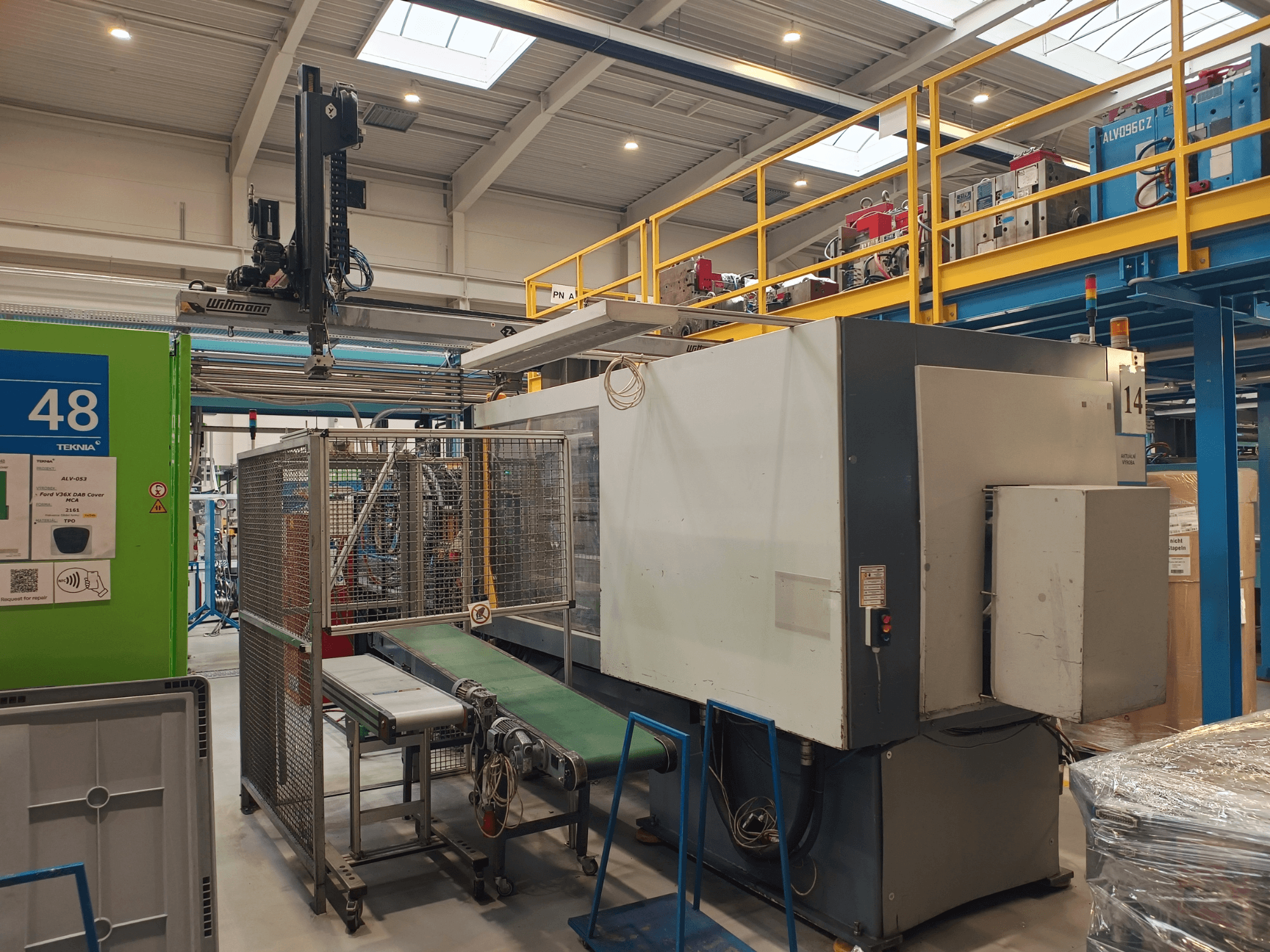 Equipo de automatización industrial con transportador verde, etiquetado "48", y puente grúa; vista lateral de la máquina en el suelo de la fábrica.